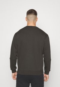 Uomo con capelli corti che indossa una felpa marrone scuro semplice e pantaloni neri, in piedi contro uno sfondo bianco, rivolto lontano dalla macchina fotografica.