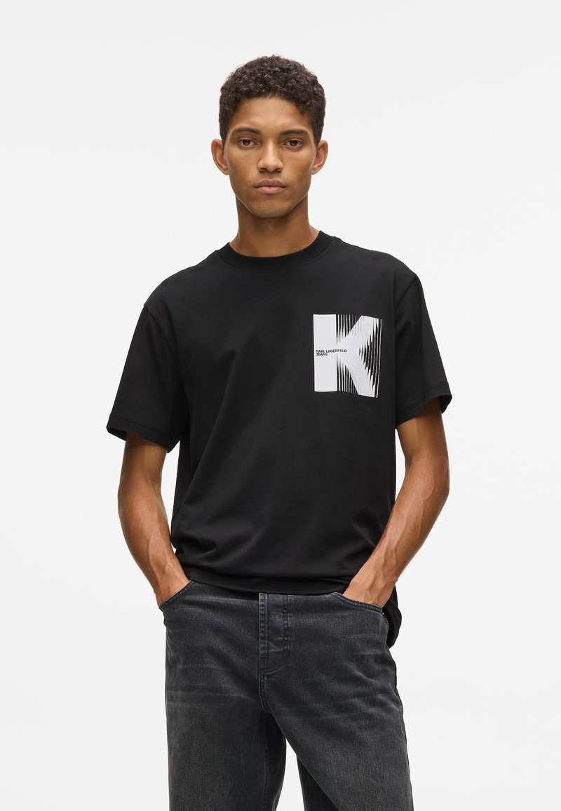 Jaunietis ar īsiem, cirtainiem matiem, ģērbies melnā T-kreklā ar lielu baltu "K" logo un tumši pelēkās džinsās, rokas makos, stāv uz baltā fona.