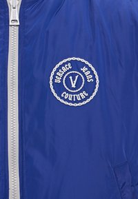 Blue bomber jacket with silver zipper, featuring a circular white embroidered logo that includes "Versace Jeans Couture" and a central "V."