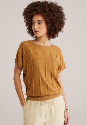 Maglia a maniche corte giallo senape in maglia con costine verticali e vestibilità ampia, abbinata a pantaloni crema con coulisse e un braccialetto d'oro.