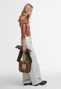 Grüne bestickte Schultertasche mit floralem Muster, kombiniert mit einem figurbetonten, langärmligen Oberteil in pink- und grün kariert, und weit geschnittenen weißen Hosen.