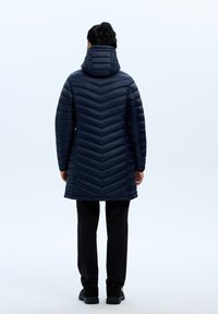 Tummansininen toppatakki, jossa on chevron-kuvio, korkea kaulus ja huppu, yhdistetty mustiin housuihin ja tummaan jalkineeseen.