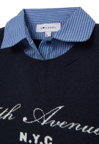 Marineblaues Sweatshirtmaterial mit weißer Schriftzug, das eine strukturierte Oberfläche aufweist. Darunter ein hellblau gestreiftes Hemd mit Kragen und Knopfverschluss.