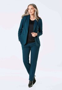 Teal blazer en bijpassende broek gemaakt van gladde stof, gecombineerd met een zwart top. De broek heeft een getailleerde pasvorm en subtiele naden.