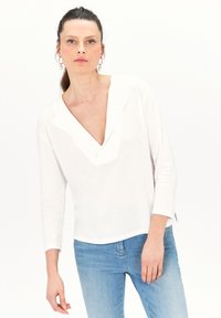 Witte lange mouw top met een V-hals en split aan de onderkant, gemaakt van zachte stof. Gecombineerd met lichtblauwe jeans, minimalistisch ontwerp.