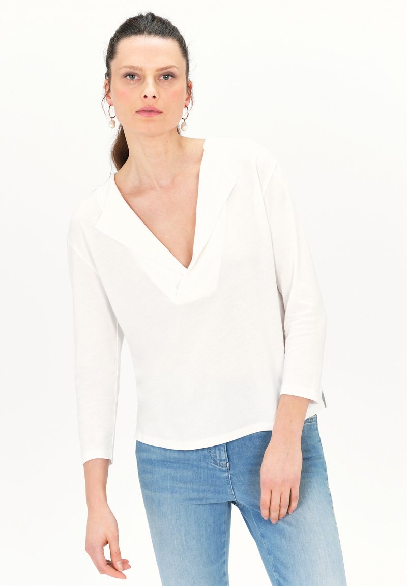 Witte lange mouw top met een V-hals en split aan de onderkant, gemaakt van zachte stof. Gecombineerd met lichtblauwe jeans, minimalistisch ontwerp.