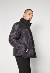 Giacca puffer camouflage nera e grigia con colletto alto, chiusura con zip e polsini elasticizzati. Tessuto liscio con sezioni trapuntate.