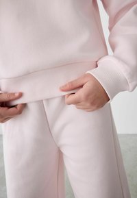 Ljusrosa sweatshirt med ribbade mudd och midja, matchande med joggers. Slät textur; fokus på plaggdetaljer och passform.