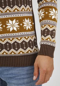 Brązowy sweter dzianinowy z białymi i musztardowymi wzorami, z motywami płatków śniegu i geometricznymi wzorami. Żebrowane mankiety i detal u dołu swetra.