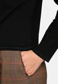 Schwarzes Langarmoberteil mit seidig-glatter Textur und einem metallisch akzentuierten Saum, kombiniert mit braunen karierten Hosen mit ausgefransten Kanten.