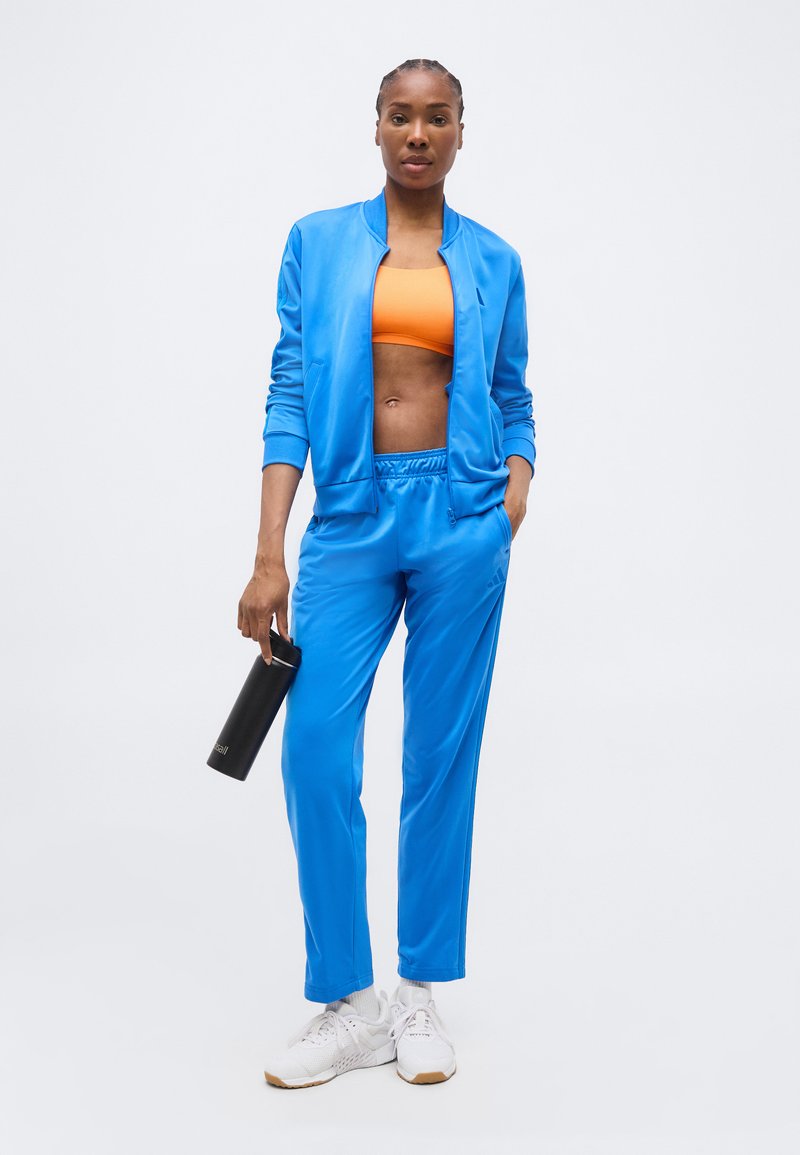 Donna in tuta blu e reggiseno sportivo arancione, che tiene una borraccia nera, indossando scarpe da ginnastica bianche su uno sfondo chiaro uniforme.