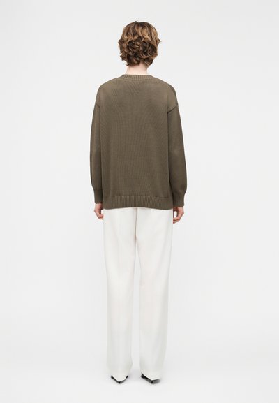 Άτομο με κοντά κυματιστά μαλλιά, φορώντας πουλόβερ χρώματος ελιάς και λευκό παντελόνι φαρδύ στο κάτω μέρος, στέκεται με την πλάτη προς τα εμπρός σε απλό λευκό φόντο.
