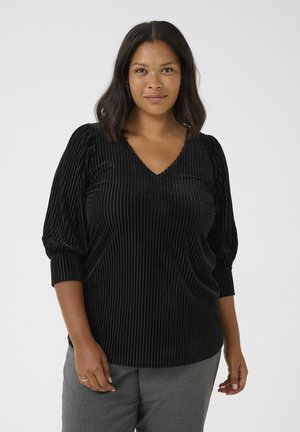 Black velvety blouse with vertical stripes, v-neckline, and puffed sleeves. The fabric has a soft texture and a relaxed fit. Paired with grey pants.