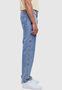 Light blue denim jeans with a straight cut, faded wash, a back pocket detail, and worn with white sneakers. Side view.