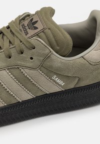 Oliivinvihreä mokkanahkainen tennari, jossa on teksturoidut oliivi- ja beige raidat, musta kumipohja ja kielen päällä oleva "adidas"-logolla varustettu merkki.