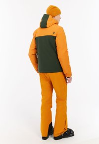 Oranžová a zelená bunda s kapucňou, ktorá má dizajn v blokoch farieb. V skombinovaní s jasne oranžovými nohavicami a čiernymi lyžiarskymi čižmami.