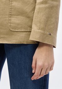 Χέρι που ακουμπά δίπλα σε μπλε τζιν, φορώντας μπεζ σακάκι με τσέπη και μικρό έμβλημα κόκκινου, λευκού και μπλε σημαίας στην μανσέτα.