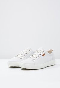 Weiße Sneaker aus glattem Leder mit runder Zehenpartie, Schnürdesign, beigen Akzenten und einer strukturierten Gummisohle. Logo-Patch an der Seite.