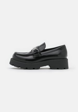 Mocasines de charol negro con punta redondeada, que cuentan con una correa frontal con hebilla y una suela gruesa y texturizada.