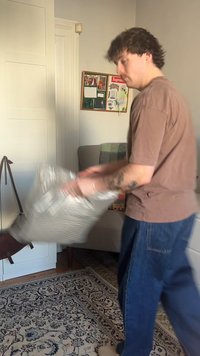 Žmogus purto pilką pagalvėlę kambaryje, kuriame yra raštuotas kilimas, sofa, sienų koliažas ir balti durys su kabančia ruda krepšiu.