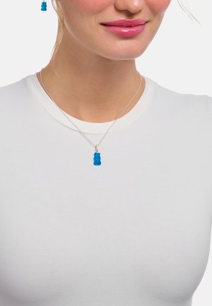 Anhänger in Form eines blauen Gummibärchens an einer dünnen silbernen Kette. Die Trägerin trägt ein schlichtes weißes Hemd, das das farbenfrohe Accessoire hervorhebt.