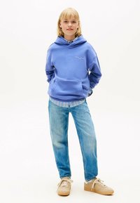 Jong persoon met blond haar, gekleed in een blauwe hoodie, gestreepte shirt, lichtblauwe jeans en beige sneakers, staand op een witte achtergrond.