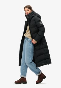 Mujer vistiendo un abrigo largo negro de plumas, suéter beige, jeans azul claro y botas marrones con cordones, de pie contra un fondo blanco.