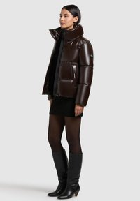 Brown padded puffer jacket with a high collar, shiny finish, and button closure. Worn over a black sweater and ribbed skirt with knee-high black boots.