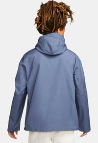 Chaqueta azul impermeable con capucha, de corte holgado, mangas largas y textura suave. Presenta un diseño sencillo en la parte posterior y acentos de botones en el dobladillo.