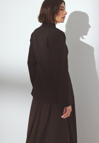 Čierny zavinovací blejzer s vysokým golierom, hladkou textúrou, priliehavým strihom a dlhými rukávmi, skombinovaný s voľnou hnedou sukňou.