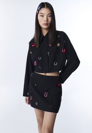 Jeune femme aux longs cheveux noirs portant une veste courte noire et une jupe ornées d'anneaux et d'épingles métalliques colorés, posant devant un fond uni.