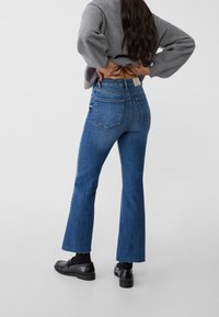 Jeans flare in denim blu con vita alta, dotati di due tasche posteriori e orlo grezzo. Abbinati a mocassini neri e un maglione grigio.