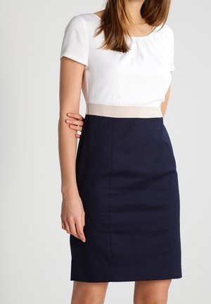 Mujer con un vestido ajustado con una parte superior blanca de manga corta, cintura beige y falda azul marino hasta la rodilla sobre un fondo liso.