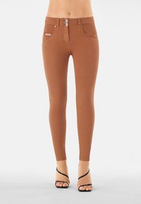 Leggings marroni con una texture liscia, design a vita alta, chiusura con doppio bottone e tasche anteriori, abbinati a tacchi neri con fasce.
