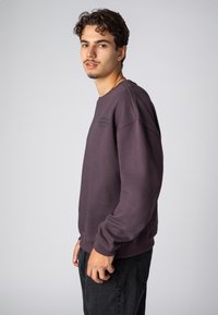 Lila Sweatshirt mit Rundhalsausschnitt, strukturiertem Stoff, leicht oversized Schnitt und schwarzem gesticktem Text auf der Brust. Kombiniert mit schwarzen Jeans.