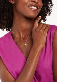 Gouden halsketting en armband met groene ovale stenen, gezet in ingewikkeld metaalwerk. Draagt een roze top, die een gladde textuur laat zien.