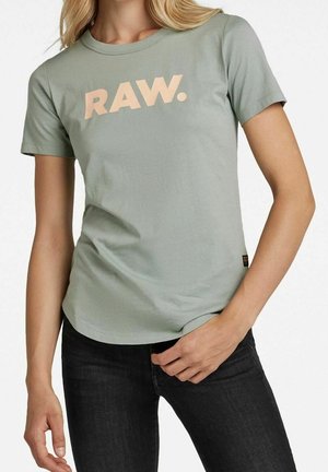 Femme portant un T-shirt à manches courtes vert clair avec le texte "RAW." en beige, associé à un jean noir, posant avec une main près de la hanche.