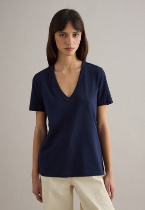 Frau mit glattem dunklem Haar, die ein marineblaues V-Ausschnitt-T-Shirt mit kurzen Ärmeln und hellbeige Hosen vor neutralem Hintergrund trägt.