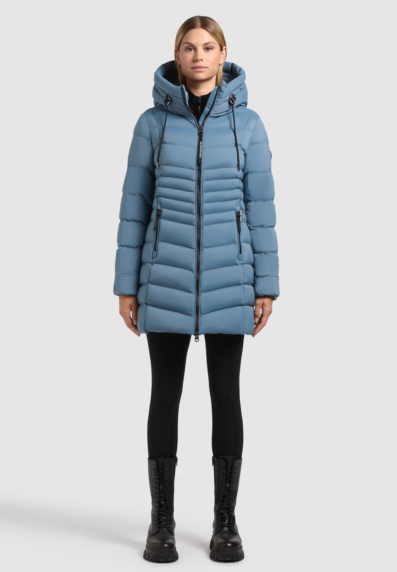 Giacca puffer blu chiaro con cappuccio, caratterizzata da cuciture diagonali, chiusura con zip e dettagli neri. Abbinata a leggings neri e stivali.