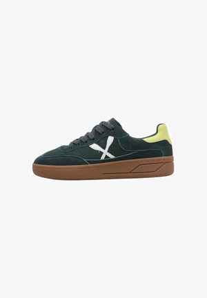 Zapatilla deportiva de ante verde oscuro con logo "X" blanco, suela marrón, cordones negros y lengüeta amarilla en el talón, mostrada de perfil.
