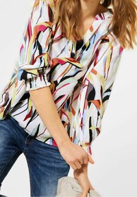 Blouse multicolore avec un motif floral aux formes allongées, col en V, et poignets élastiqués. Le matériau semble léger et texturé.