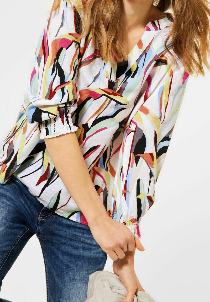 Blouse multicolore avec un motif floral aux formes allongées, col en V, et poignets élastiqués. Le matériau semble léger et texturé.