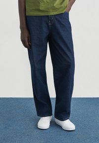 Σκούρα μπλε τζιν παντελόνια με σχέδιο ίσιου ποδιού, φορεμένα με πράσινο πουκάμισο και λευκά αθλητικά παπούτσια. Διαθέτουν εμφανείς ραφές και τσέπες.