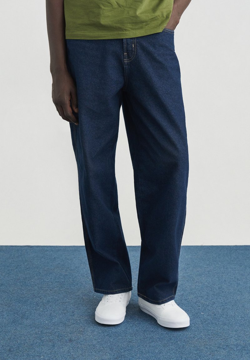 Σκούρα μπλε τζιν παντελόνια με σχέδιο ίσιου ποδιού, φορεμένα με πράσινο πουκάμισο και λευκά αθλητικά παπούτσια. Διαθέτουν εμφανείς ραφές και τσέπες.