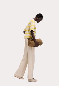 Pruhovaná košile s krátkým rukávem v žluté a béžové barvě, široké krémové kalhoty, držící hnědou semišovou kabelku, stojící na jednobarevném pozadí.