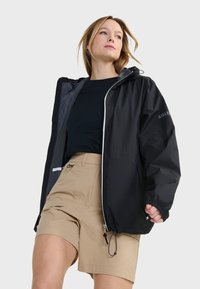 Veste noire imperméable avec fermeture éclair à l'avant, capuche ajustable et poignets élastiques, associée à un short beige avec plusieurs poches.