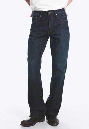 Personne portant un jean en denim bleu foncé et des bottes en cuir noir, debout devant un fond uni.