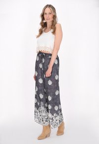 Witte gecropte tanktop met kantafwerking; grijze wijde broek met een patroon en witte bloemenontwerpen; gedragen met lichtbruine enkellaarzen.