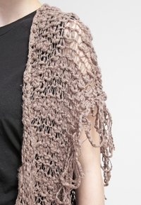 Ljusbrun stickad sjal med en lös, öppen väv och fransar, draperad över en svart topp, som visar textur och lager.