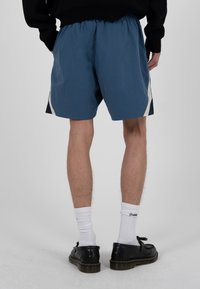Mann trägt blaue sportliche Shorts mit weißen und schwarzen Seitenstreifen, weiße Socken und schwarze Lederschuhe, steht auf einem grauen Boden.